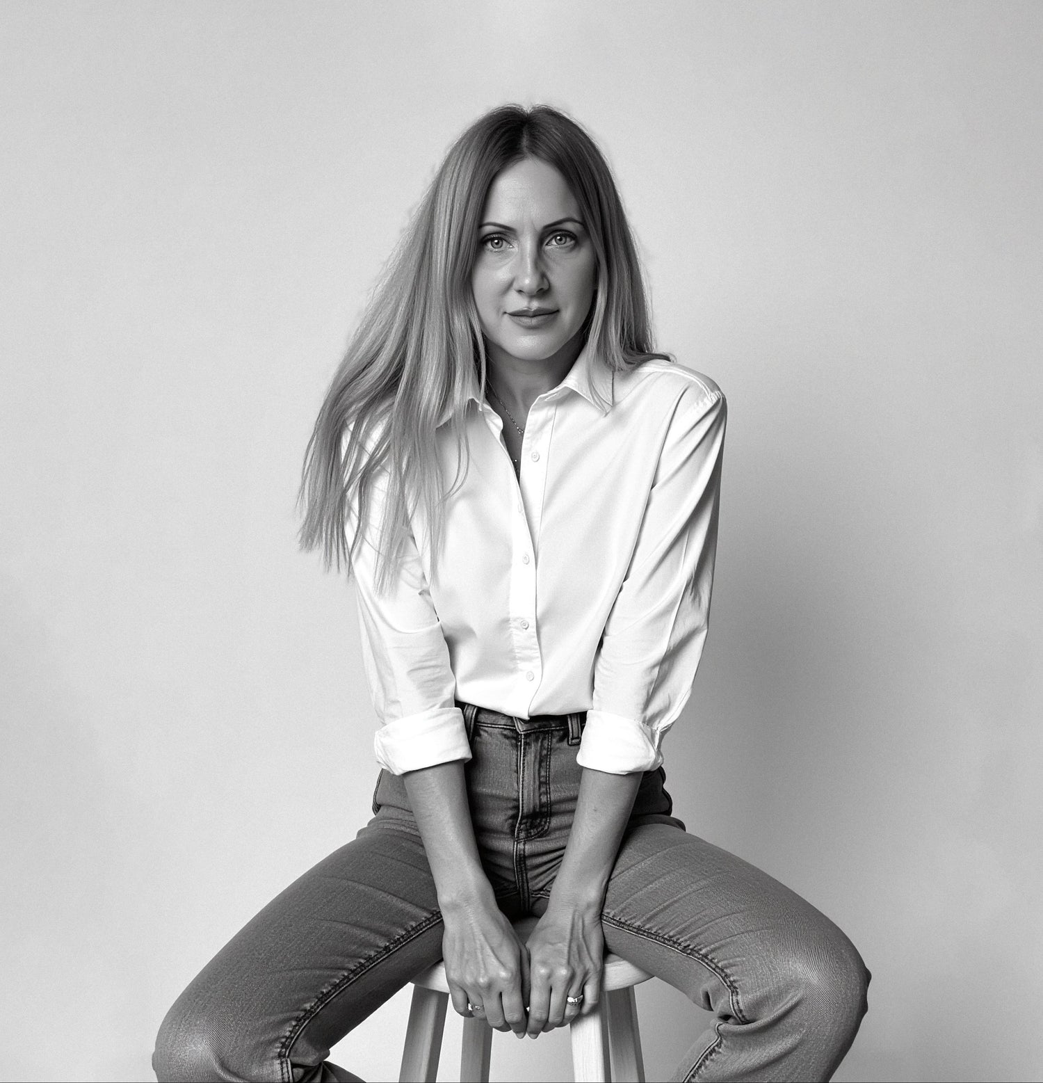 Noemi Safir the international artist. Founder of NOE MI ART COUTURE in Black and white portrait sitting on a stool against a plain background